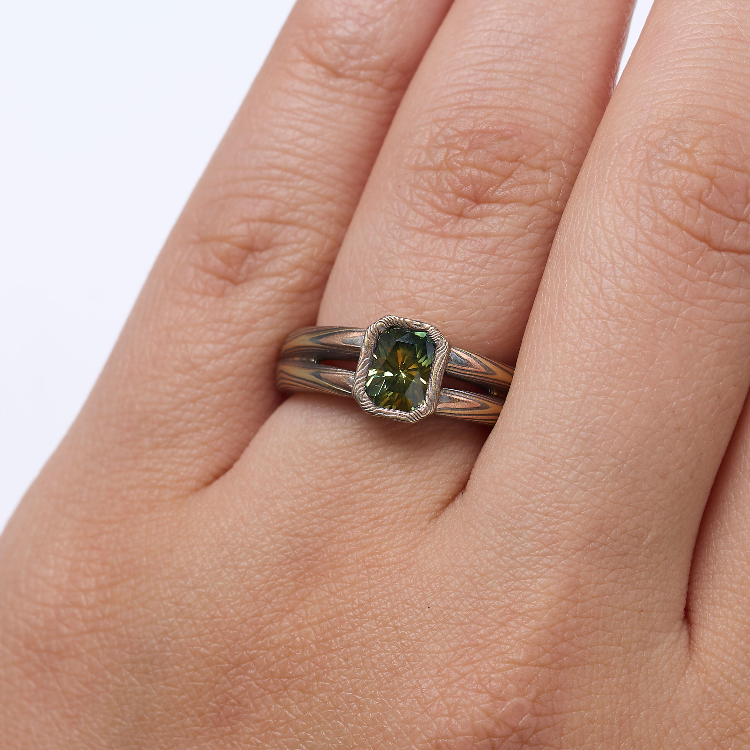 Mokume gane engagement ring with a split shank tapered towards the back of the ring, and a single green emerald cut sapphire placed at the front, with a rim of patterned metal around it.